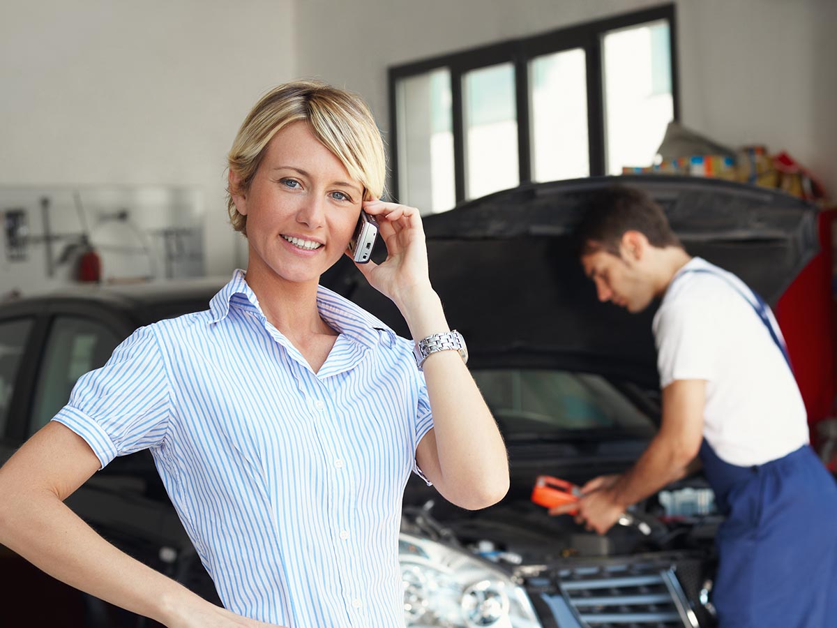 Smiling woman on the phone while a mechanic works on her car